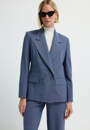 Blazer doppiopetto in tessuto a scacchi blu chiaro, con revers a punta e due tasche frontali. Indossato con un dolcevita crema sotto.