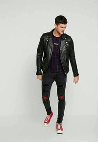 Νέος άνδρας περπατάει, φοράει μαύρο δερμάτινο μπουφάν, ρίγες σκούρου χρώματος μπλούζα, σκισμένα μαύρα τζιν με κόκκινες λεπτομέρειες και κόκκινα ψηλά αθλητικά παπούτσια.