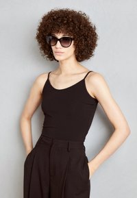 Black fitted tank top with thin straps, worn with high-waisted black trousers. Model has curly hair and sunglasses, against a gray background.