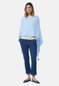 Kobieta stojąca z rękami w kieszeniach, ubrana w jasnoniebieski szal na kremowym swetrze, ciemnoniebieskie dżinsy 3/4 oraz niebieskie czółenka.