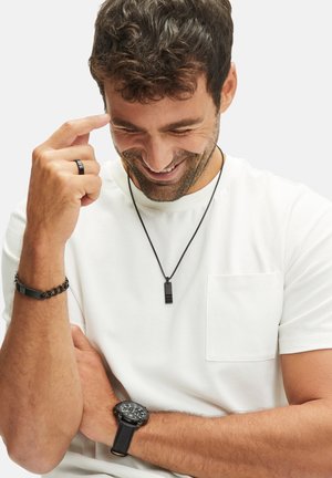 Zwarte hanger ketting op een dunne koord, zwarte textuur armband, zwart horloge met een ronde wijzerplaat, gedragen over een wit t-shirt met een borstzak.