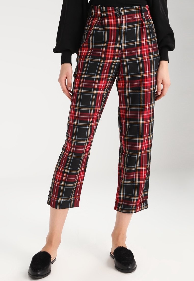 Femme portant un pantalon court à carreaux rouge et noir avec une ceinture noire boutonnée, associé à des chaussures noires sans lacets avec pompons.