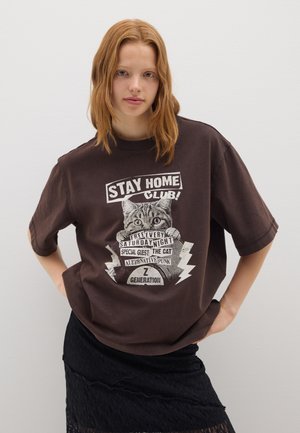 Camiseta de gran tamaño color marrón con una impresión gráfica que presenta un gato y elementos de texto. Mangas cortas, cuello redondo, tejido de algodón suave.