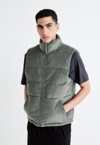 Vesta puffer din catifea verde, cu guler înalt, fermoar frontal și buzunare laterale. Pur tată pe un tricou negru cu mânecă scurtă, are un material texturat.