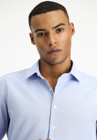 Camicia azzurra chiaro con motivo a piccoli quadretti, colletto a punta, taschino sul petto e trama liscia. Presenta bottoni bianchi.