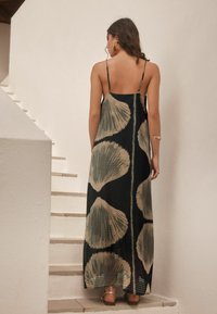 Robe maxi noire avec un motif de coquillage beige, dotée de fines bretelles et d'un tissu texturé. Portée par un mannequin sur des escaliers.