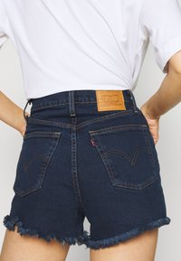 Shorts en denim foncé avec un ourlet effiloché, présentant une étiquette en cuir marron à l'arrière, et des poches arrière traditionnelles avec des coutures contrastées.