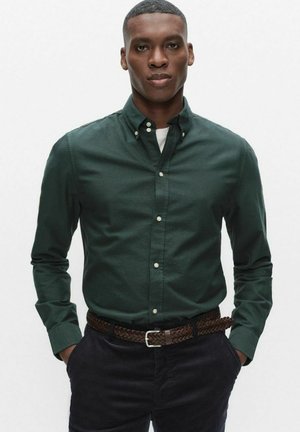 Man met een donkere huid, gekleed in een donkergroen overhemd met knopen, zwarte broek en een bruine gevlochten riem, staand met handen in de zakken.