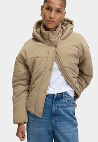 Beige pufferjakke med hætte, der har lynlås og trykknaplukning, quiltet tekstur og en afslappet pasform, båret over en hvid skjorte og blå jeans.