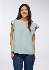 Blusa blu teal chiaro con maniche corte a balze, tessuto morbido, scollatura rotonda e design frontale plissettato, abbinata a jeans scuri.