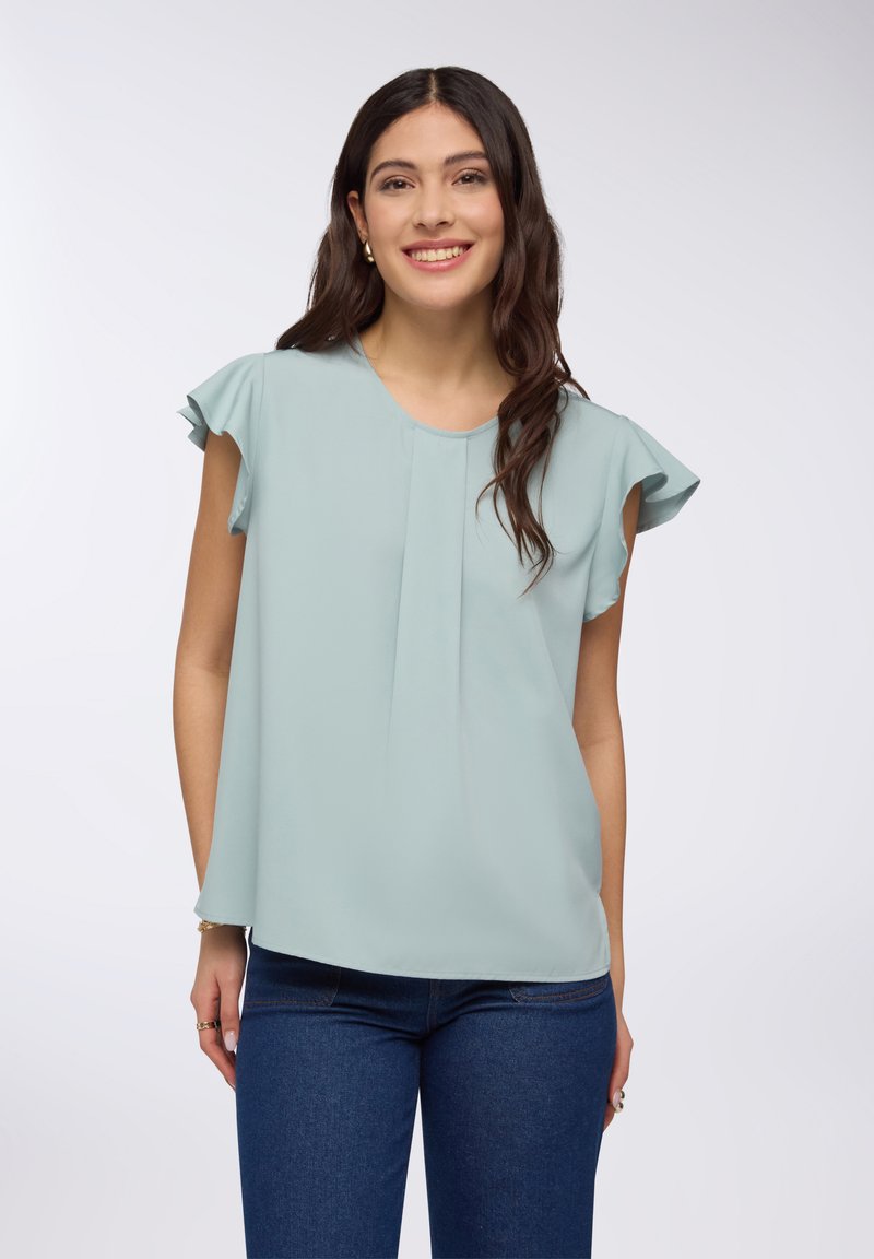 Blusa blu teal chiaro con maniche corte a balze, tessuto morbido, scollatura rotonda e design frontale plissettato, abbinata a jeans scuri.