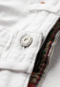 Overol de algodón blanco con un botón plateado etiquetado "Superdry Denim". El forro interior presenta un colorido patrón floral con costuras contrastantes.