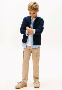 Marinblå stickad kofta över en ljusrandig skjorta, kombinerad med beige cargobyxor och beige sneakers, med en avslappnad passform och framfickor.