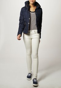 Marineblaues Puffermantel mit Kapuze, gestreiftes schwarz-weißes Oberteil, weiße Skinny-Jeans und blaue Sneakers mit weißen Schnürsenkeln.