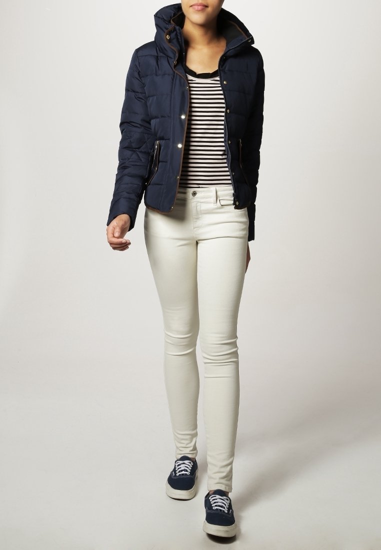Marineblaues Puffermantel mit Kapuze, gestreiftes schwarz-weißes Oberteil, weiße Skinny-Jeans und blaue Sneakers mit weißen Schnürsenkeln.