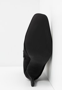Chaussure noire à talon, positionnée côté semelle vers le haut contre un coin blanc, montrant une semelle lisse et un talon aiguille étroit.
