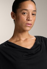 Crna majica s cowl izrezom, koja ima mekanu teksturu. Tkanina se elegantno obavija oko ramena, stvarajući glatku siluetu.