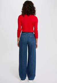Maglietta rossa a coste con maniche lunghe abbinata a jeans a gamba larga blu in denim a vita alta, dotati di tasche posteriori e sneakers bianche.