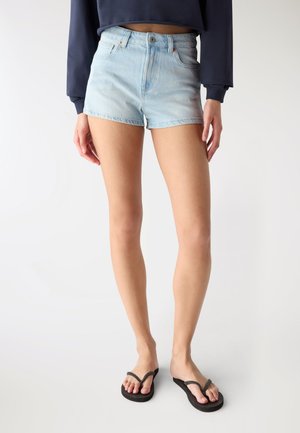 Donna con pantaloncini di jeans azzurri chiari, top corto a maniche lunghe blu navy e sandali infradito neri, in piedi davanti a uno sfondo semplice.