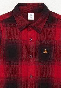 Κόκκινο και μαύρο καρό πουκάμισο φλις με γιακά, διαθέτει καφέ κουμπιά και μια μικρή κεντημένη αρκούδα στην τσέπη του στήθους.