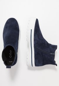 Námořnické modré semišové kotníkové boty s bílou gumovou podrážkou, bočním zipem, polstrovaným límcem a kontrastním modrým pruhem podél podrážky.