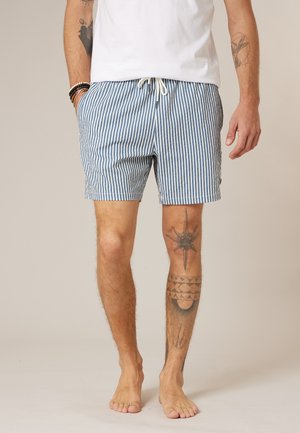 Homme portant un short rayé vertical bleu et blanc avec des poches avant et un cordon blanc, un t-shirt blanc, et plusieurs tatouages sur les jambes et les bras.