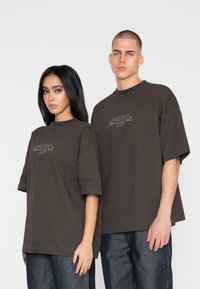 Brune oversized t-shirts med korte ærmer og et logo i midten. Modellerne bærer mørke denimbund, hvilket fremhæver en afslappet stil.