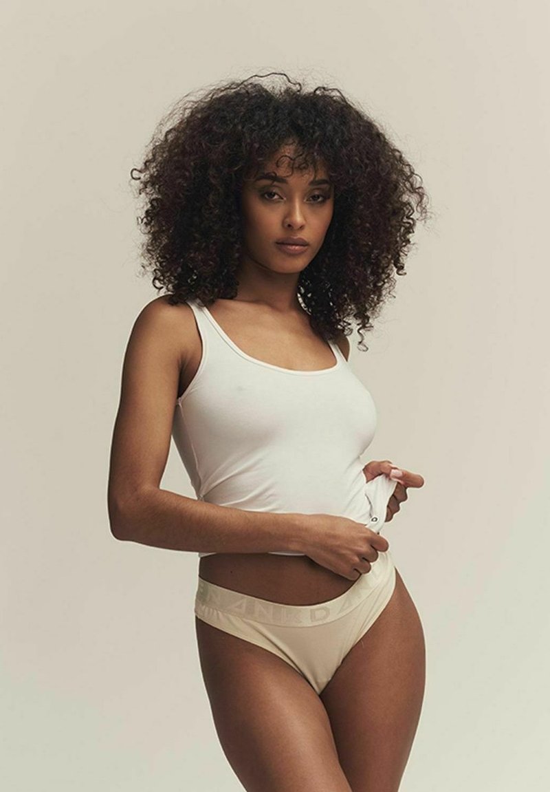 Un débardeur blanc et un sous-vêtement beige clair avec une ceinture de marque. Le modèle a les cheveux bouclés et pose en ajustant ses vêtements d'une main.