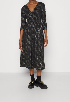 Schwarzes Langarmkleid mit Wickeldesign, das ein goldgesprenkeltes Muster und eine taillierte Passform aufweist, kombiniert mit schwarzen Plateauschuhen.