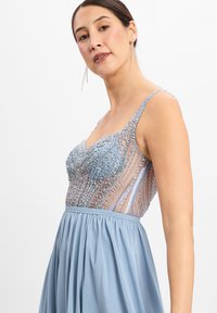Hellblaues Abendkleid mit einem bestickten, transparenten Oberteil und einem fließenden, plissierten Rock. Es hat dünne Träger und aufwendige Verzierungen.