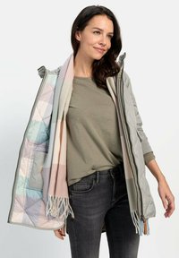 Gepatchte graue Jacke mit einem pastellfarbenen Karo-Futter; über einem grünen Hemd getragen; ergänzt durch einen fransigen Schal in sanften Tönen, kombiniert mit dunklen Jeans.