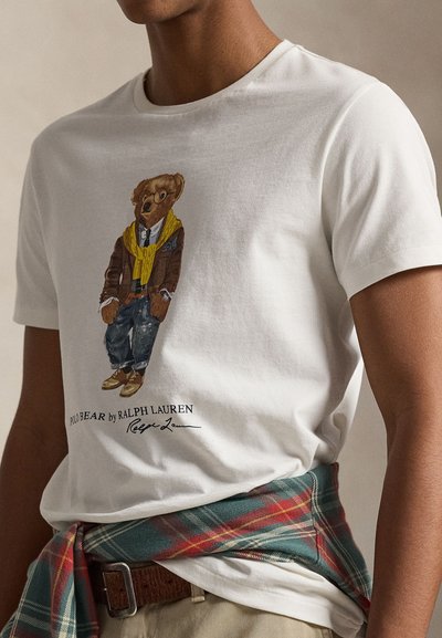 Tricou alb din bumbac cu o ilustrație a unui urs îmbrăcat într-un sacou maro, eșarfă galbenă, blugi albaștri și pantofi maro; logo și semnătură dedesubt.