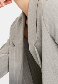 Blazer gris clair avec rayures verticales fines, revers structurés et texture de tissu lisse. Poche accentuée visible sur le côté gauche.