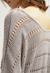 Pull en tricot gris clair avec des motifs ajourés, doté d'un large col et d'un détail en chaîne argentée décoratif le long de la couture de l'épaule.
