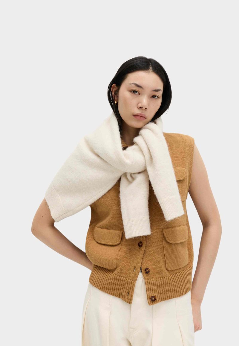 Junge Frau mit kurzen schwarzen Haaren, die eine ärmellose braune Strickweste trägt und einen weißen Pullover über die Schultern gelegt hat.
