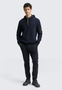 Navy capuchon sweatshirt met een voorkantzak, gouden trekkoord en zijzak met rits. Gecombineerd met donkere tapered broek en zwarte sneakers.
