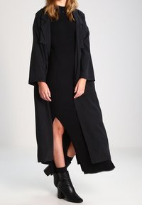 Schwarzer langer Mantel über einem figurbetonten schwarzen Rippenkleid mit seitlichem Schlitz, kombiniert mit schwarzen, hohen Stiefeletten. Glatte Textur, minimalistisches Design.