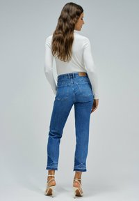 Jeans de mezclilla en azul medio, de pierna recta con un dobladillo recortado. Los bolsillos traseros tienen un bordado sutil. Combinados con una blusa de manga larga blanca.