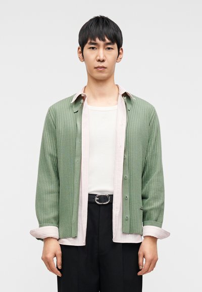 Νεαρός άνδρας που φοράει στρωτό πράσινο ζακέτα, ανοιχτό ροζ πουκάμισο, λευκό μπλουζάκι και μαύρο παντελόνι με ζώνη, στέκεται μπροστά σε απλό φόντο.