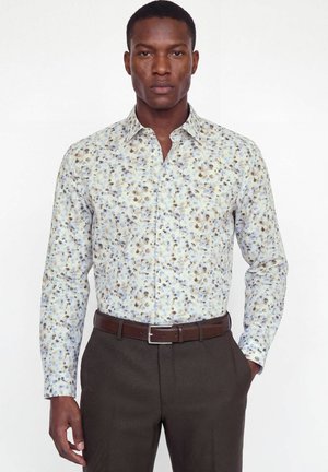 Homme portant une chemise à manches longues boutonnée avec un motif floral bleu et marron, ceinture marron et pantalon marron foncé, mains dans les poches.