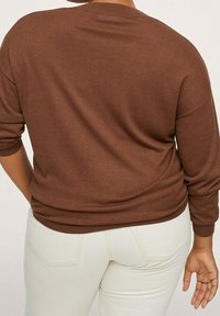 Pull en maille marron à manches longues, coupe décontractée, avec un ourlet arrondi, associé à un jean de couleur claire. Texture lisse, design simple.