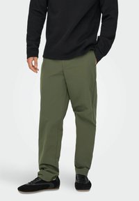 Olivgrüne Hose mit entspannter Passform, aus leichtem Material, mit seitlichen Taschen und geradem Bein, kombiniert mit schwarzen Schuhen.