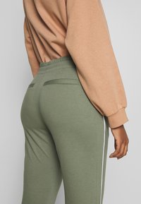 Olivgrüne Jogginghosen mit elastischem Bund, ausgestattet mit zwei Gesäßtaschen und weißen Seitenstreifen, kombiniert mit einem hellbraunen Sweatshirt.