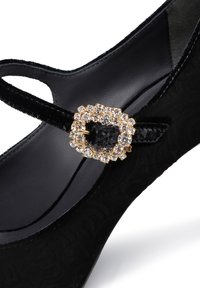 Scarpa in velluto nero con una delicata fascetta dotata di una fibbia in tono oro decorata con cristalli trasparenti. Superficie testurizzata e finitura morbida.