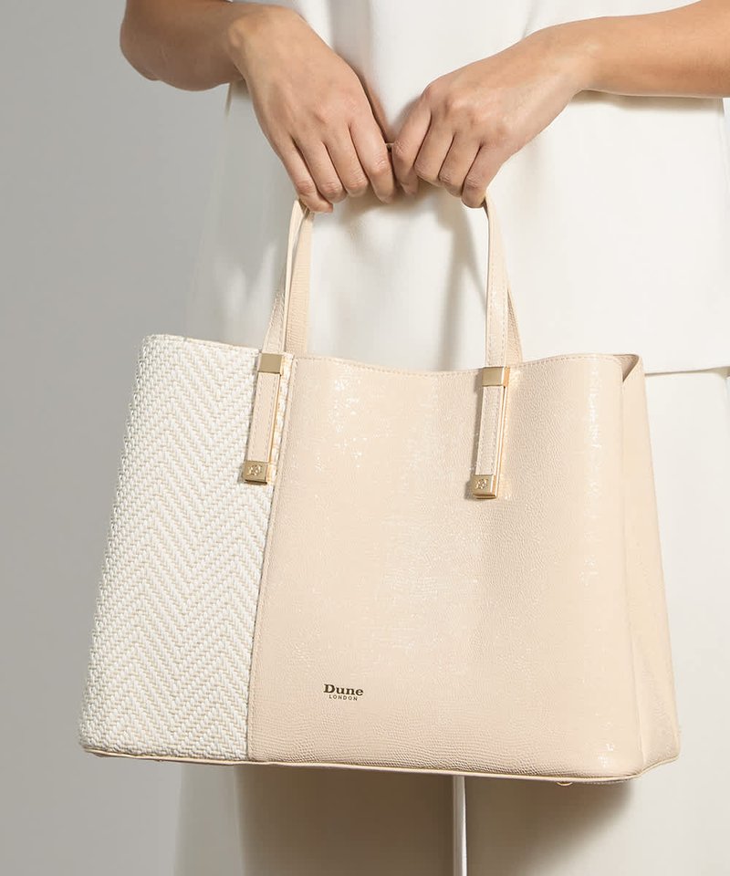 Des mains tiennent un sac cabas beige et blanc Dune London avec deux anses, présentant une texture tissée sur un côté.