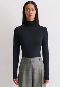 Frau steht in einem schwarzen langärmeligen Rollkragenpullover, der in hoch geschnittene graue weite Hosen gesteckt ist, vor einem schlichten weißen Hintergrund.