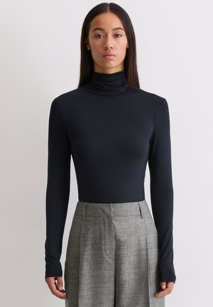 Vrouw staande in een zwarte langwerpige coltrui, gestopt in een grijze hoge taille wijde broek, tegen een effen witte achtergrond.