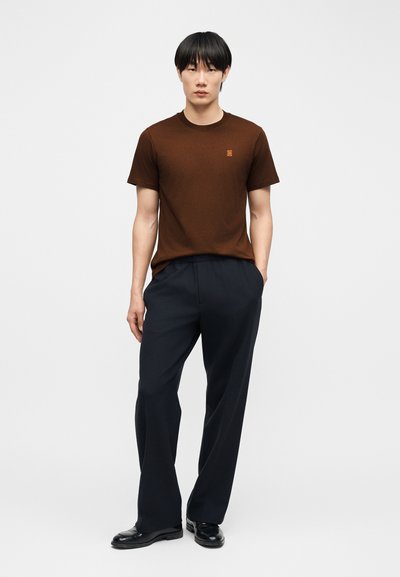 Jeune homme portant un T-shirt marron à manches courtes, un pantalon noir à jambes larges et des chaussures noires, debout avec une main dans la poche.