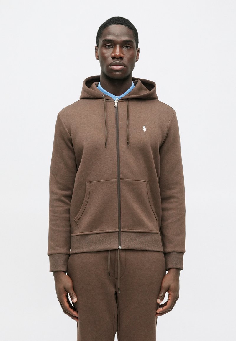 Smeđa zip-up dukserica od mekog materijala, s prednjim džepom, podesivim špagama i malim logotipom na lijevoj strani prsiju.