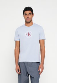 Jasnoniebieski bawełniany t-shirt z okrągłym dekoltem, kr�ótkimi rękawami i nadrukiem na przodzie z napisem „Calvin Klein” oraz „cK” w czerwono-białych kolorach.